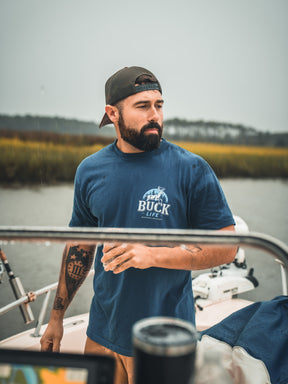 A bearded man in a blue Mucker Bucket Buck Life Premium Tee and backwards cap stands on a boat holding a drink, with marshland and water in the background.