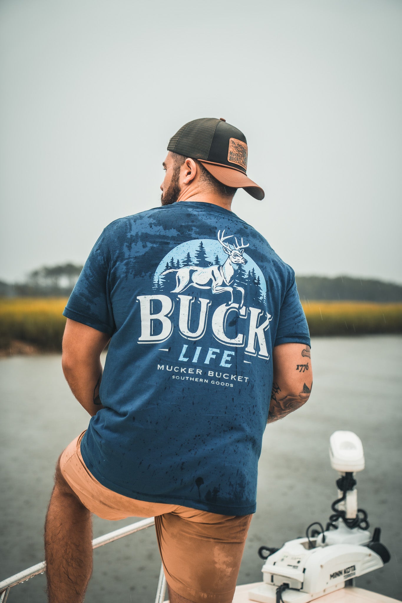 A man in a blue Buck Life Premium Tee by Mucker Bucket, with a backward cap, sits on a boat looking over a river. The premium relaxed fit shirt features a deer and trees graphic for a calm, outdoorsy vibe.