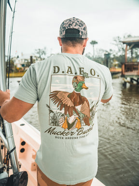 A man on a boat holds a fishing rod while wearing the DAFO Premium Tee by Mucker Bucket, featuring an illustrated duck and Duck Around Find Out. Water and trees can be seen in the background.