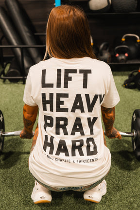 A tattooed individual squats with a barbell, wearing the Lift Heavy. Pray Hard. Tee by Tango Charlie Apparel. The T-shirt’s bold slogan reflects strength and faith, with gym equipment visible in the background.