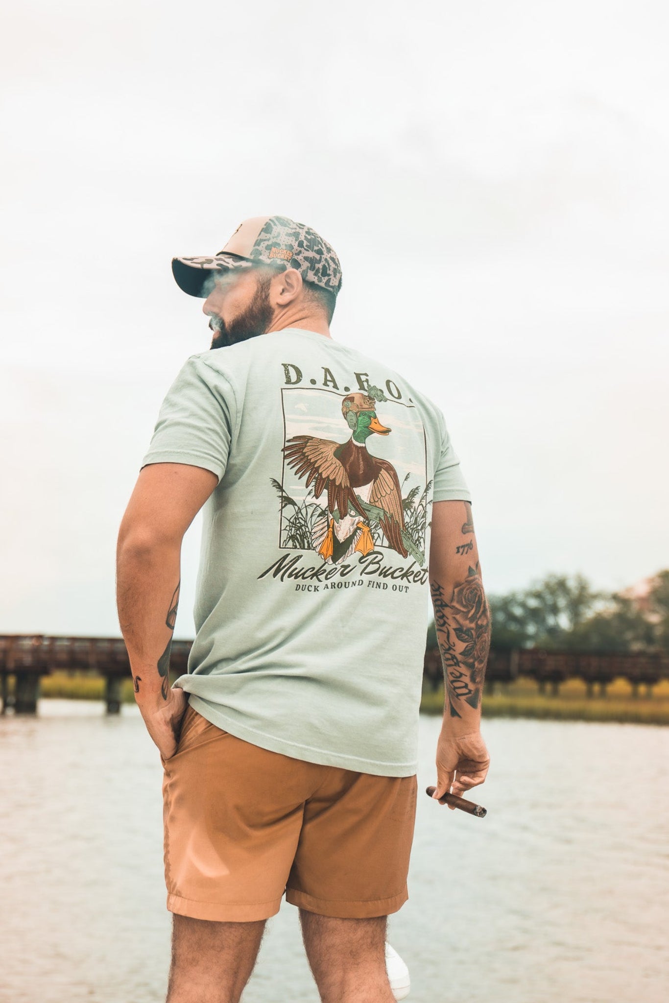 A man in a cap, Mucker Bucket DAFO Premium Tee, and shorts stands by the water holding a cigar, facing away with trees, a dock, and cloudy sky in the background.