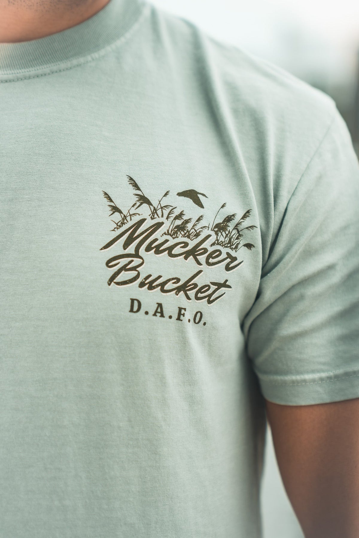 A person wearing the Mucker Bucket DAFO Premium Tee, a relaxed fit light green shirt with a garment-dyed finish and a palm tree, bird, and “Mucker Bucket D.A.F.O.” graphic on the left chest.