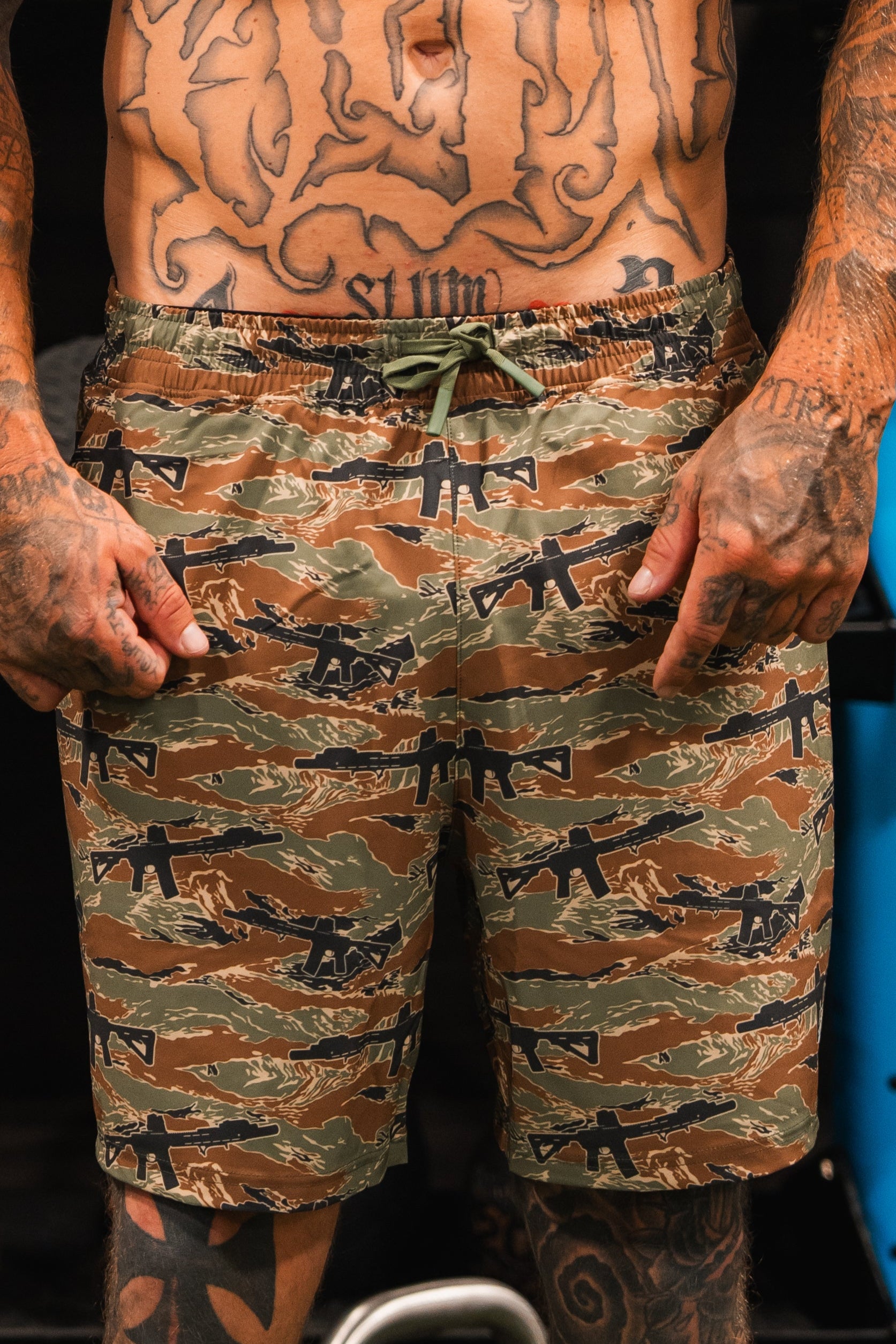 A heavily tattooed person stands indoors, pointing at their Tango Charlie Apparel Men's Performance Training Shorts | 5.5"- Snipes & Stripes—versatile hybrid shorts ideal for gym-to-swim activities.