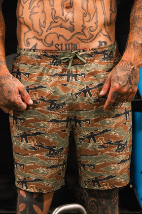 A heavily tattooed person stands indoors, pointing at their Tango Charlie Apparel Men's Performance Training Shorts | 5.5"- Snipes & Stripes—versatile hybrid shorts ideal for gym-to-swim activities.