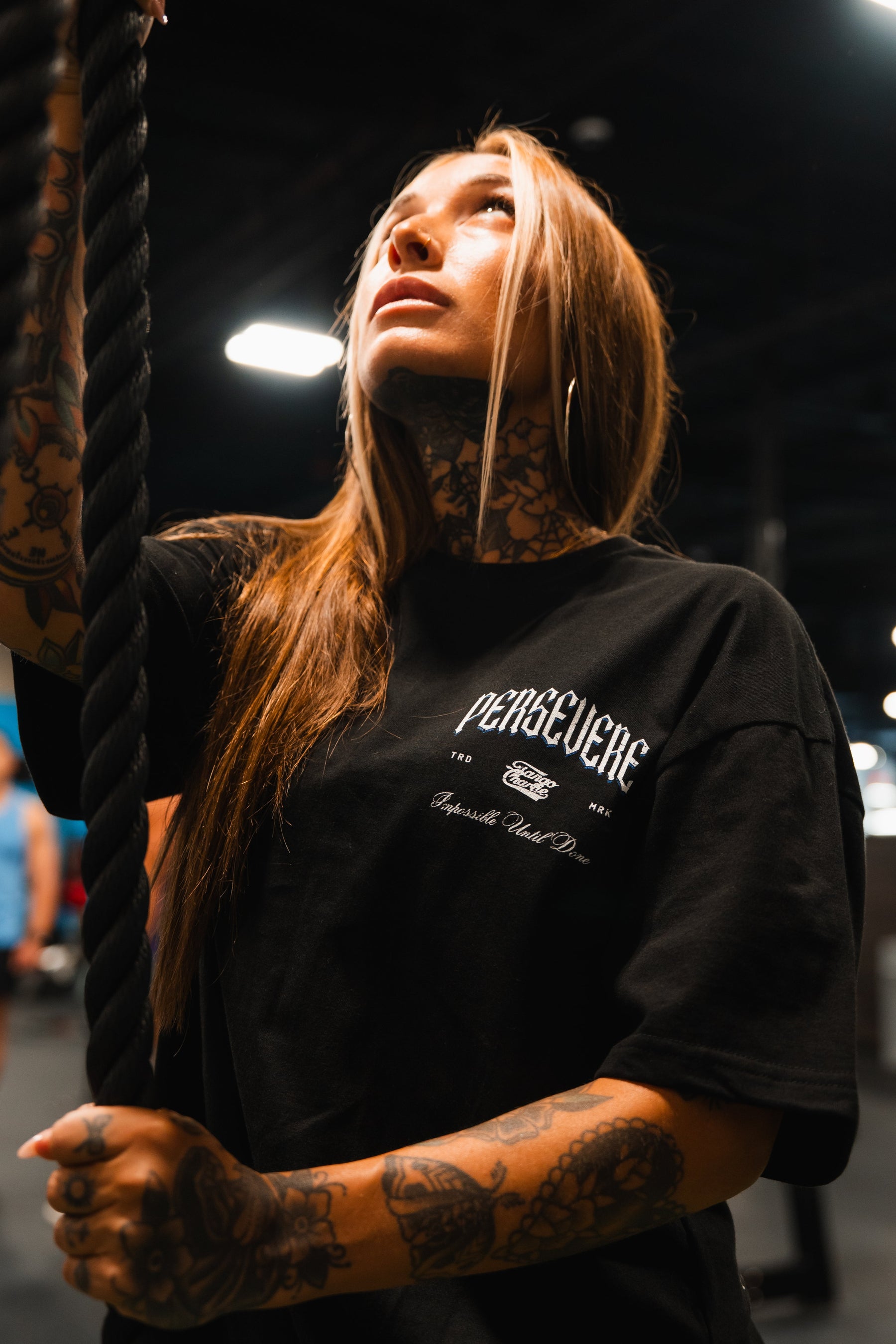 A tattooed woman with long hair holds a rope and looks up in a brightly lit gym, wearing the Tango Charlie Apparel Persevere Premium Oversized Tee—a unisex black shirt with white "PERSEVERE" text, crafted from heavyweight cotton.