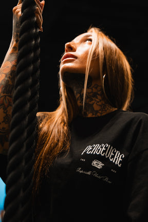 A tattooed woman grips a thick rope and looks up with determination, spotlighting the "PERSEVERE" message on a Persevere Premium Oversized Tee by Tango Charlie Apparel amid a dark backdrop.