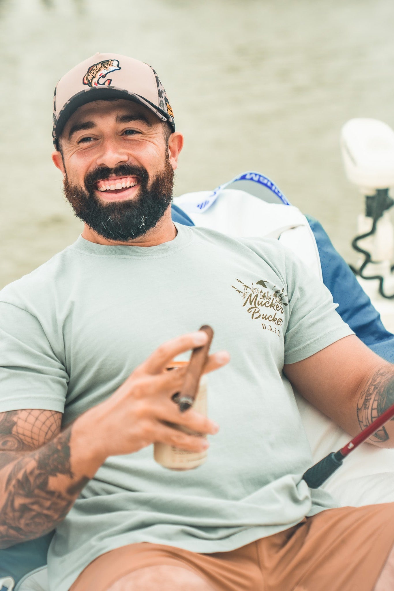 A smiling bearded man with tattoos relaxes on a boat, holding a fishing rod and cigar. He wears the Mucker Bucket DAFO Premium Tee in light blue and a beige cap with fish logo, with water in the background.