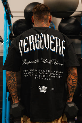 A tattooed athlete stands in the gym with dumbbells, wearing Tango Charlie Apparel’s Persevere Premium Oversized Tee, which features “PERSEVERE—Impossible Until Done—Continue in a course of action even in the face of difficulty.”.