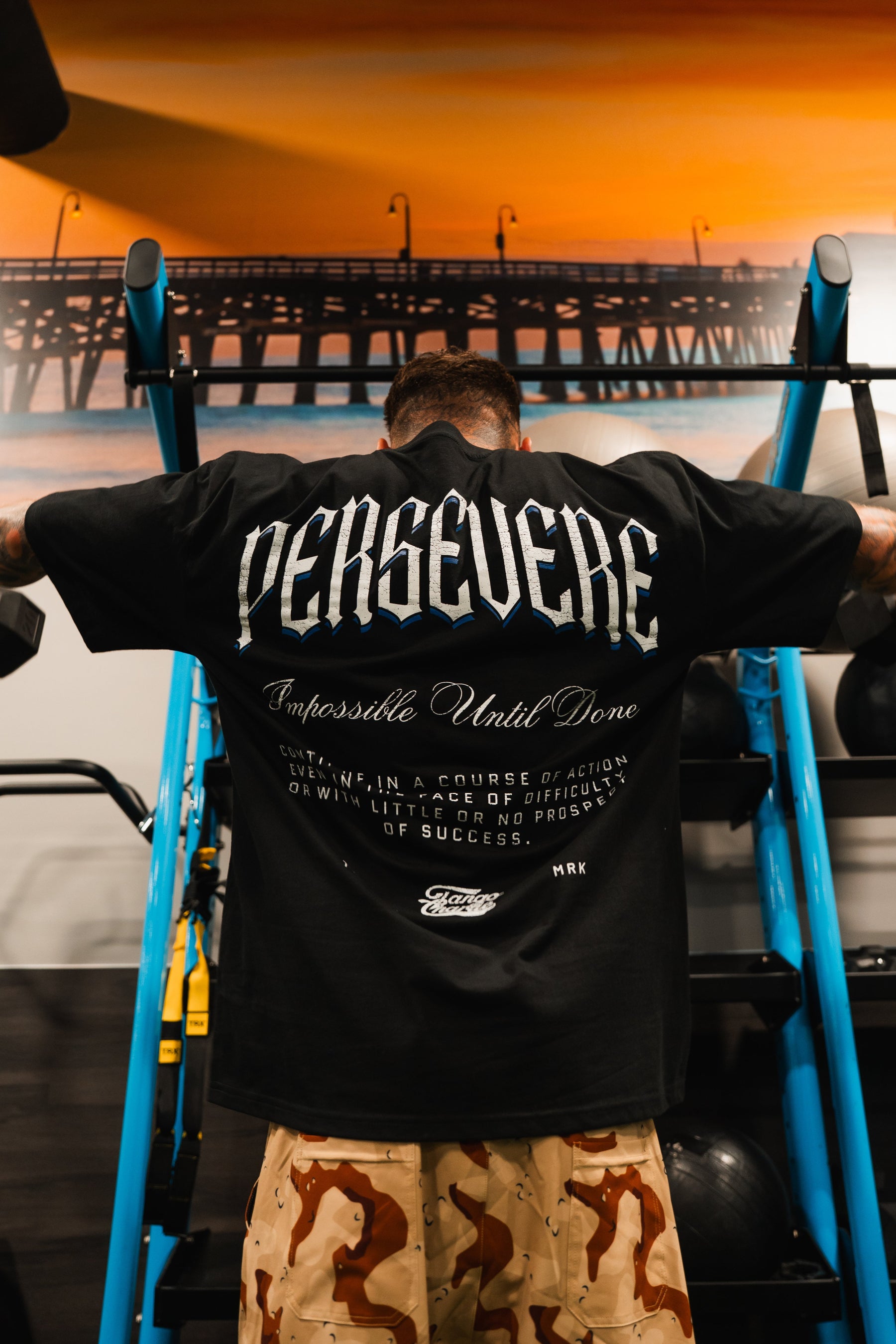 A person in the Tango Charlie Apparel Persevere Premium Oversized Tee with "PERSEVERE" boldly on the back stands by gym equipment, motivational text below, camo pants, and a pier photo hanging on the wall.