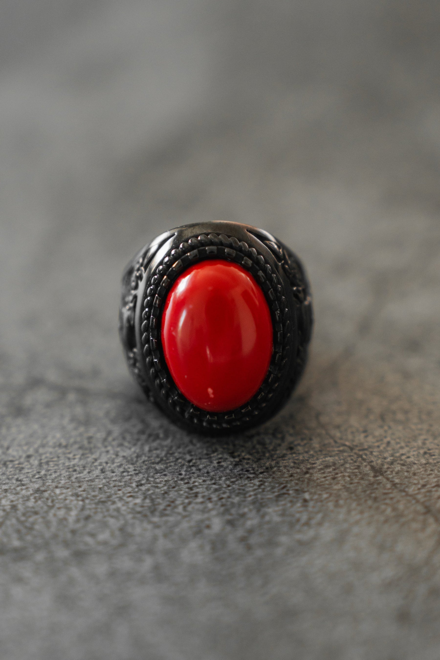 A close-up of the Relentless Betrayal Burning Rose Blackout Ring, a stainless steel piece with an ornate band and large oval red stone, is shown on a gray textured surface.