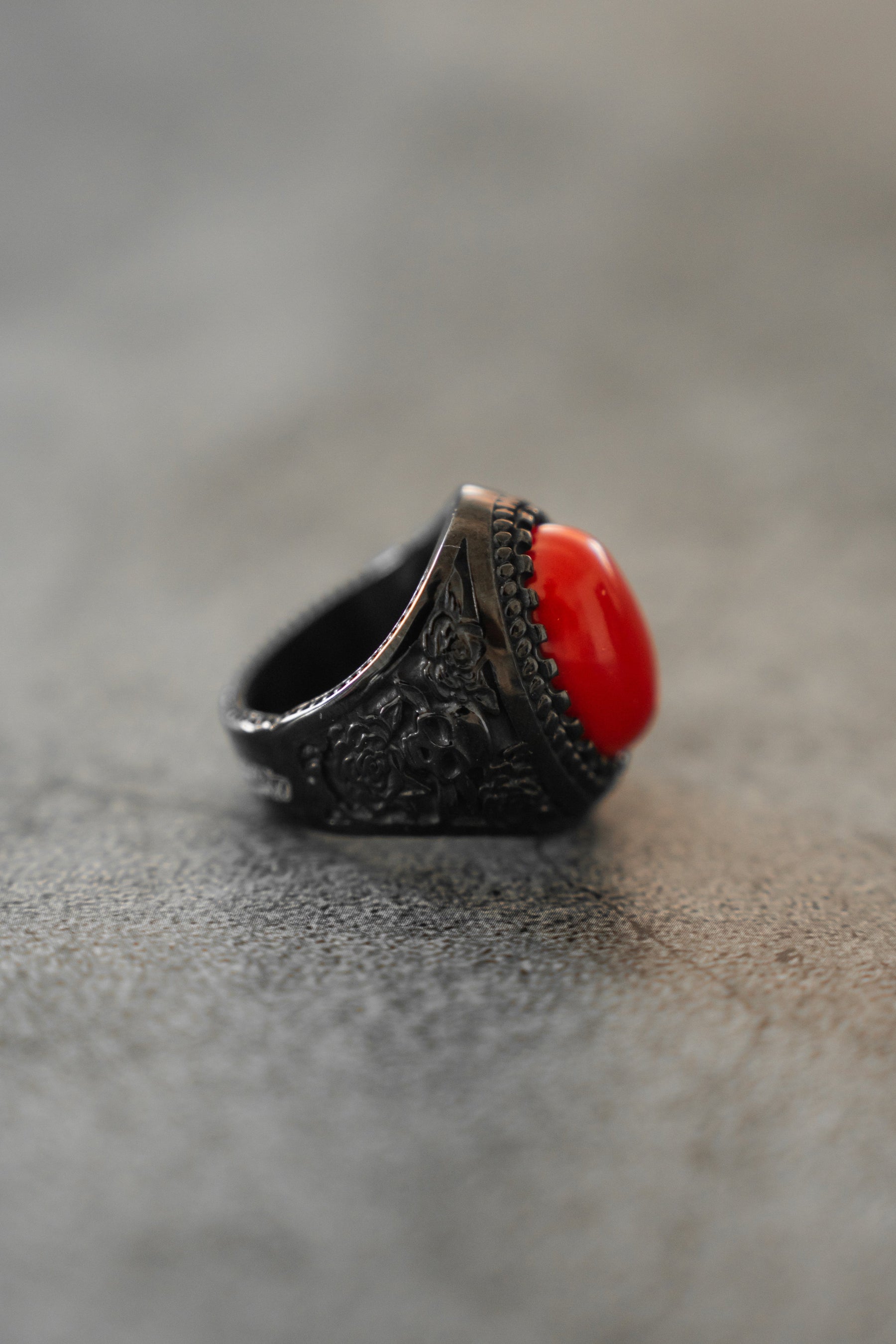 The Relentless Betrayal Burning Rose Blackout Ring, with detailed floral engravings and an oval red gemstone, is showcased on a gray textured surface.
