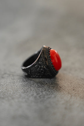 The Relentless Betrayal Burning Rose Blackout Ring, with detailed floral engravings and an oval red gemstone, is showcased on a gray textured surface.