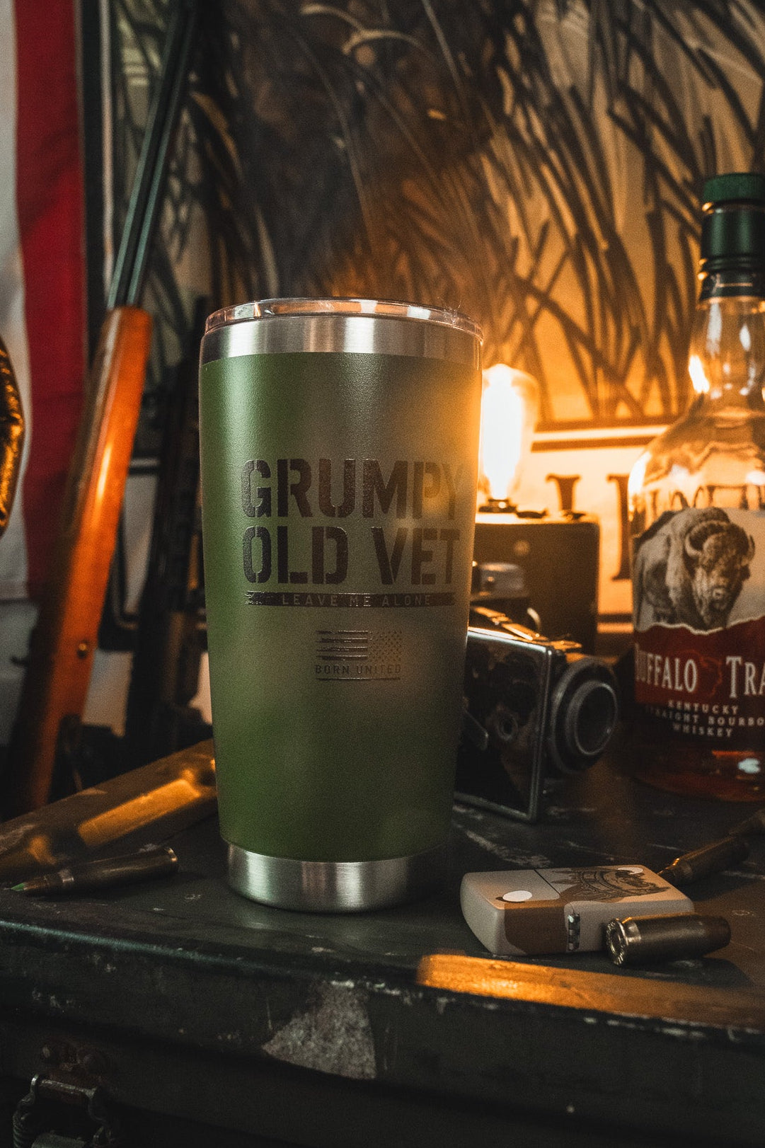 The Born United Private Label Grumpy Old Vet Tumbler - 20oz sits on a cluttered table with a whiskey bottle, camera, bullets, and lighter, as a warm lamp highlights an American flag and rifle in the background.
