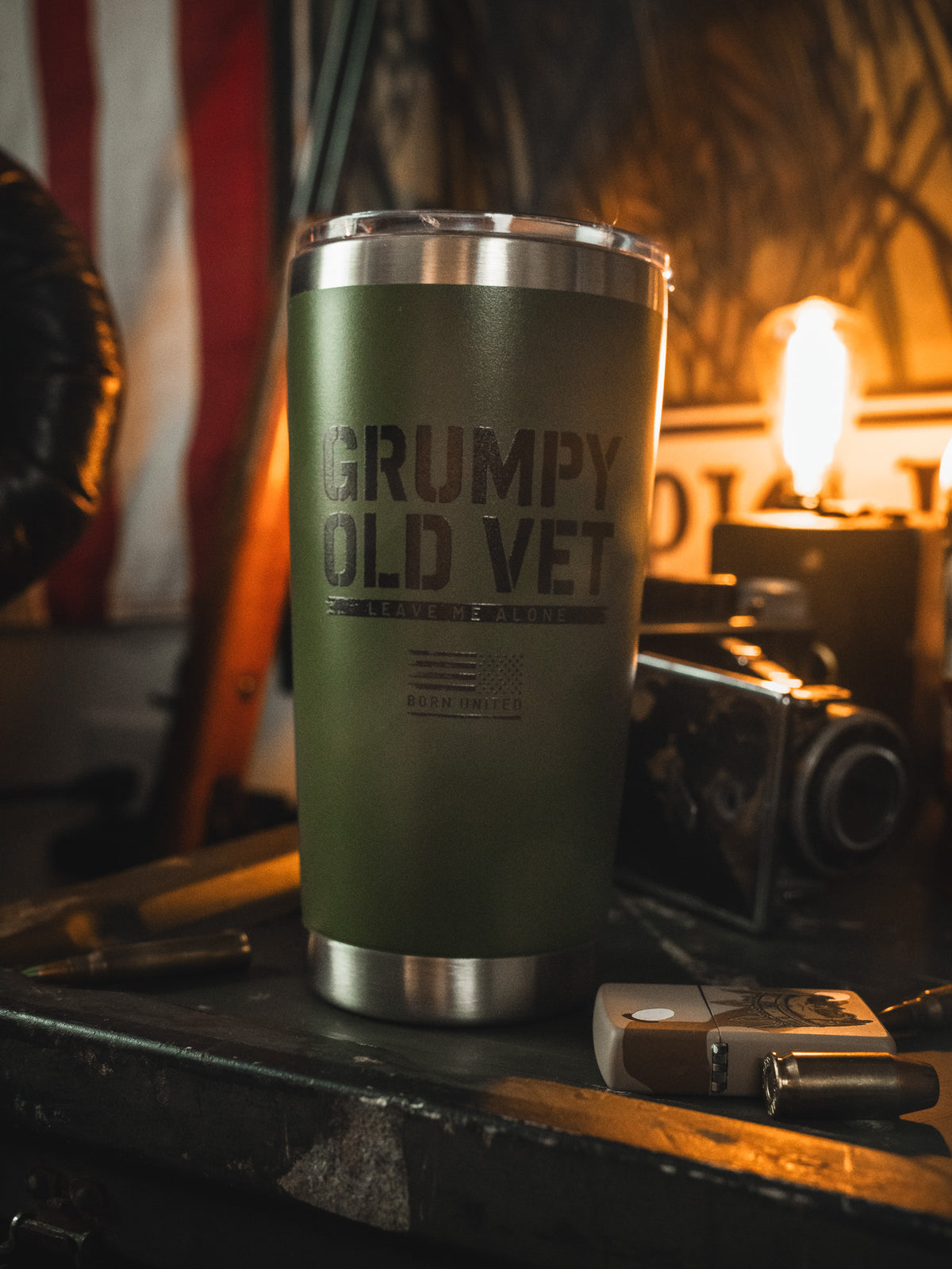 A Born United Private Label 20oz Grumpy Old Vet Tumbler in green, featuring the printed phrase, rests among vintage items on a cluttered surface with a glowing bulb in the background.