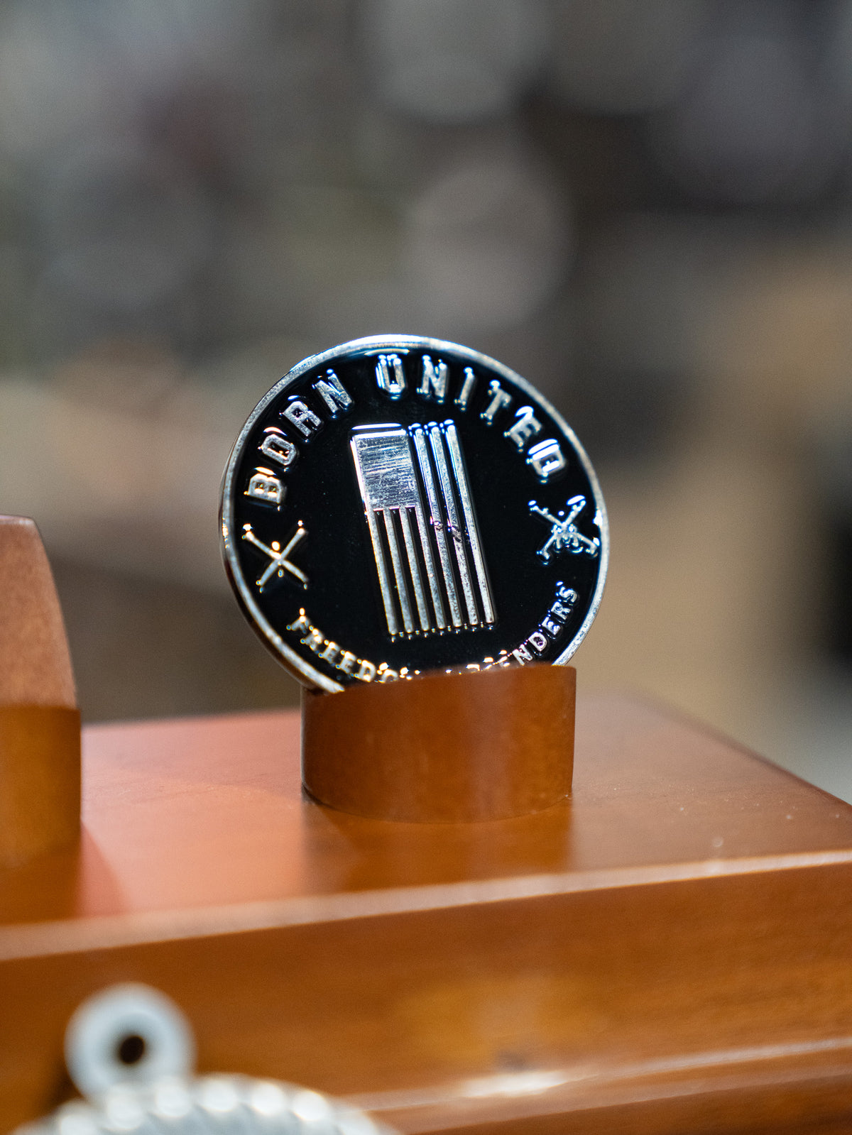 The Buzz N' Woody Challenge Coin by Born United Private Label is a round metallic coin with vertical bars, crossed swords, and ODAN UNITAS HONOR AND VICTORY around the edge, displayed on a wooden stand like collectible memorabilia.