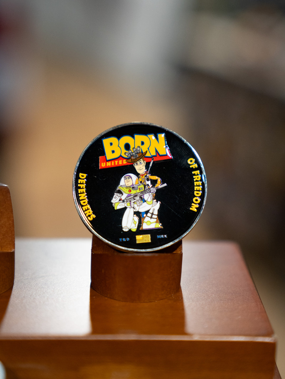 The Buzz N' Woody Challenge Coin by Born United Private Label features a soldier with a gun and American flag, "Born Defenders of Freedom" text, and is displayed upright on a wooden stand—ideal for fans of heroic spirit.