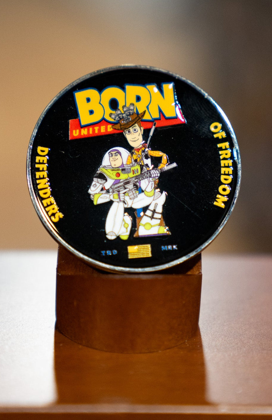 The Buzz N' Woody Challenge Coin by Born United Private Label features a soldier with a gun and American flag, "Born Defenders of Freedom" text, and is displayed upright on a wooden stand—ideal for fans of heroic spirit.