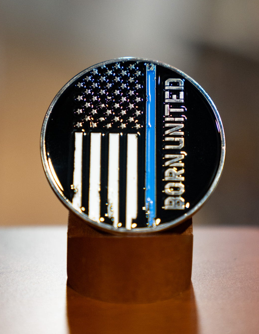 The Blue Line Flag Challenge Coin by Born United Private Label features a black-and-white American flag with a blue stripe and "Born United" along the right, displayed upright on a brown stand.
