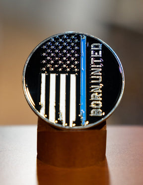 The Blue Line Flag Challenge Coin by Born United Private Label features a black-and-white American flag with a blue stripe and "Born United" along the right, displayed upright on a brown stand.