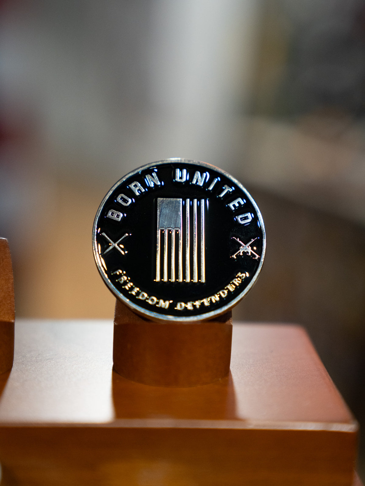 Display the Blue Line Flag Challenge Coin by Born United Private Label—a round metal coin with a U.S. flag, crossed rifles, and the words BORN UNITED and FREEDOM DIVIDED—upright on its wooden stand to show your support.