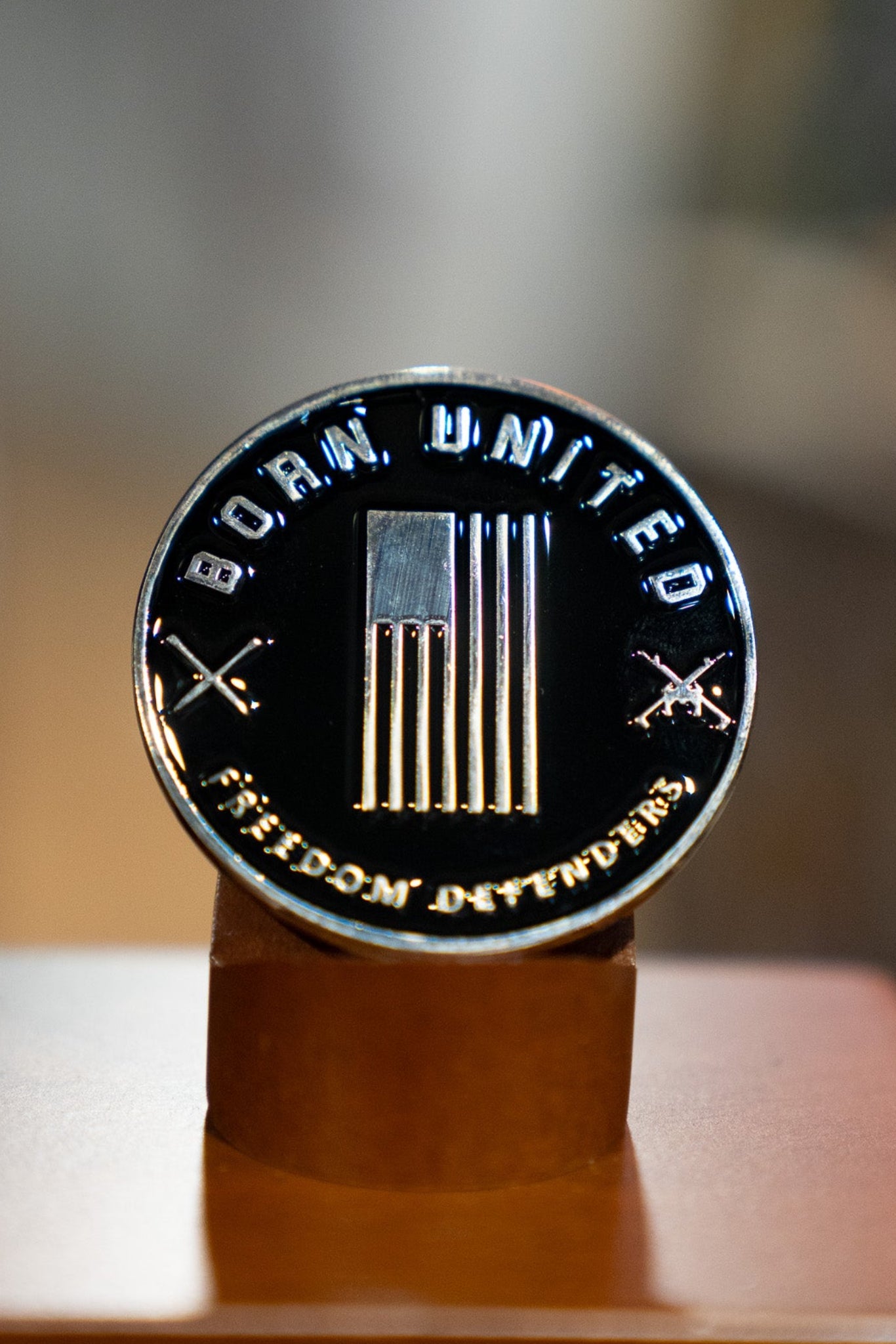 Display the Blue Line Flag Challenge Coin by Born United Private Label—a round metal coin with a U.S. flag, crossed rifles, and the words BORN UNITED and FREEDOM DIVIDED—upright on its wooden stand to show your support.