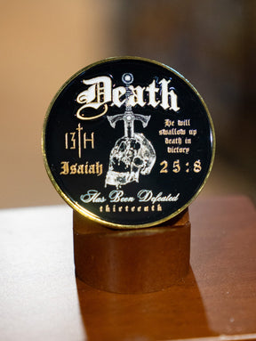 The 13th Death's Defeat Challenge Coin is a round black and gold coin featuring a skull, sword, and the inscription "He will swallow up death in victory, Isaiah 25:8," displayed upright.