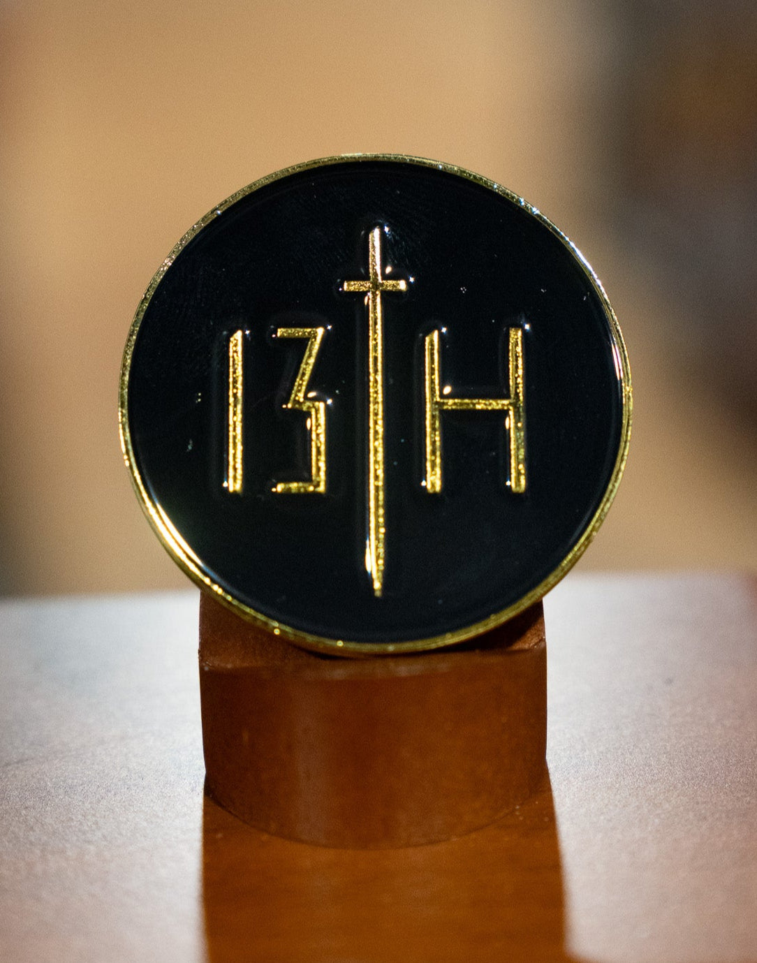 The 13th Death's Defeat Challenge Coin is a round black coin with gold edges, featuring the number 13 and letter H split by a vertical gold sword, propped upright on a brown stand.