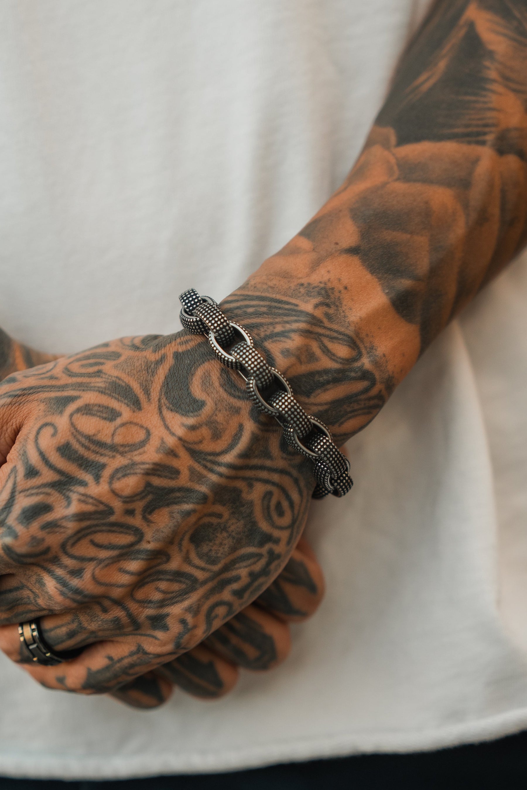 A tattooed hand and forearm wear a Relentless Betrayal Seeker Bracelet in black and silver chain plus a black ring. The person is dressed in a white shirt.