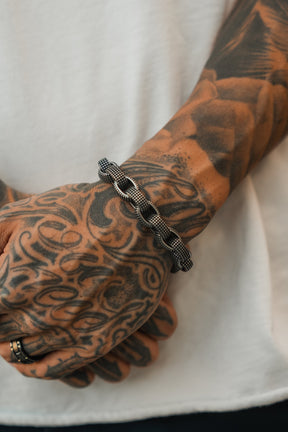 A tattooed hand and forearm wear a Relentless Betrayal Seeker Bracelet in black and silver chain plus a black ring. The person is dressed in a white shirt.