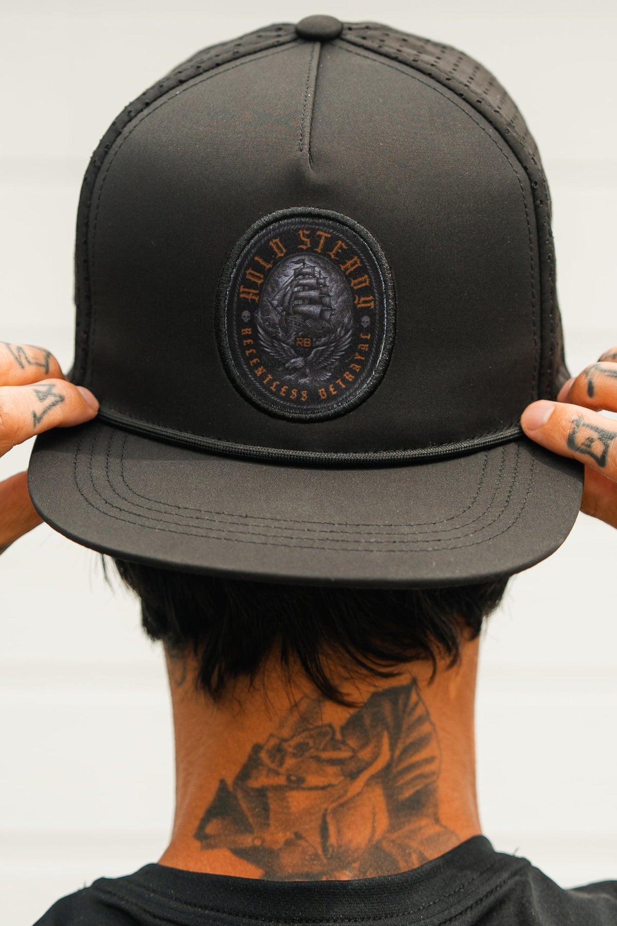 A person, seen from behind, adjusts a Relentless Betrayal Hold Steady Ghost Snapback featuring a circular patch. Tattoos are visible on their fingers and neck against a plain, light background.