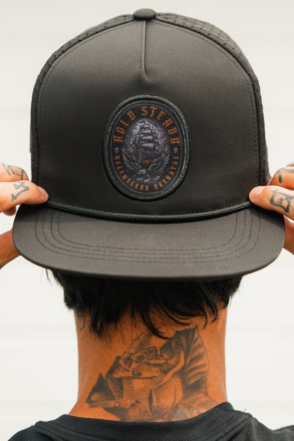 A tattooed individual adjusts a Relentless Betrayal Hold Steady Ghost Snapback, featuring a skull and text patch. Their face is hidden as the photo is taken from behind.