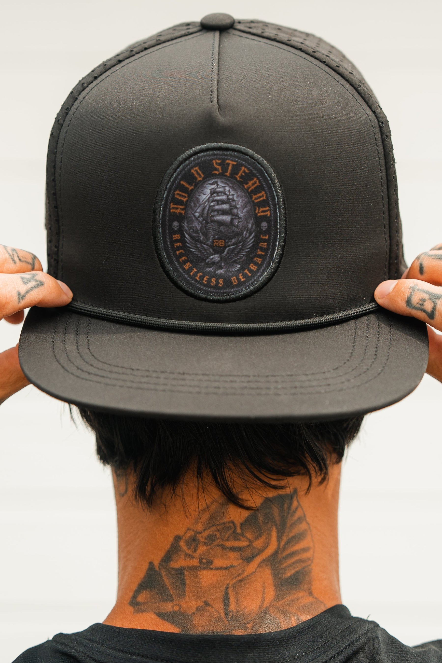 A tattooed individual adjusts a Relentless Betrayal Hold Steady Ghost Snapback, featuring a skull and text patch. Their face is hidden as the photo is taken from behind.