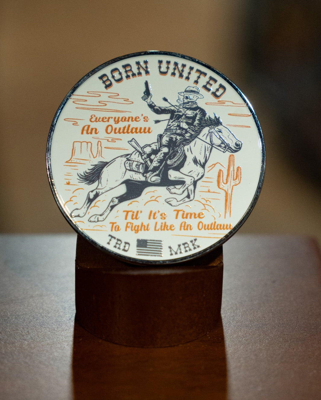 The Outlaw Challenge Coin by Born United Private Label is a round metal coin featuring a cowboy on a bucking horse, two cacti, and the text: BORN UNITED. Everyone's an outlaw until it’s time to fight like an outlaw. TRD MRK.