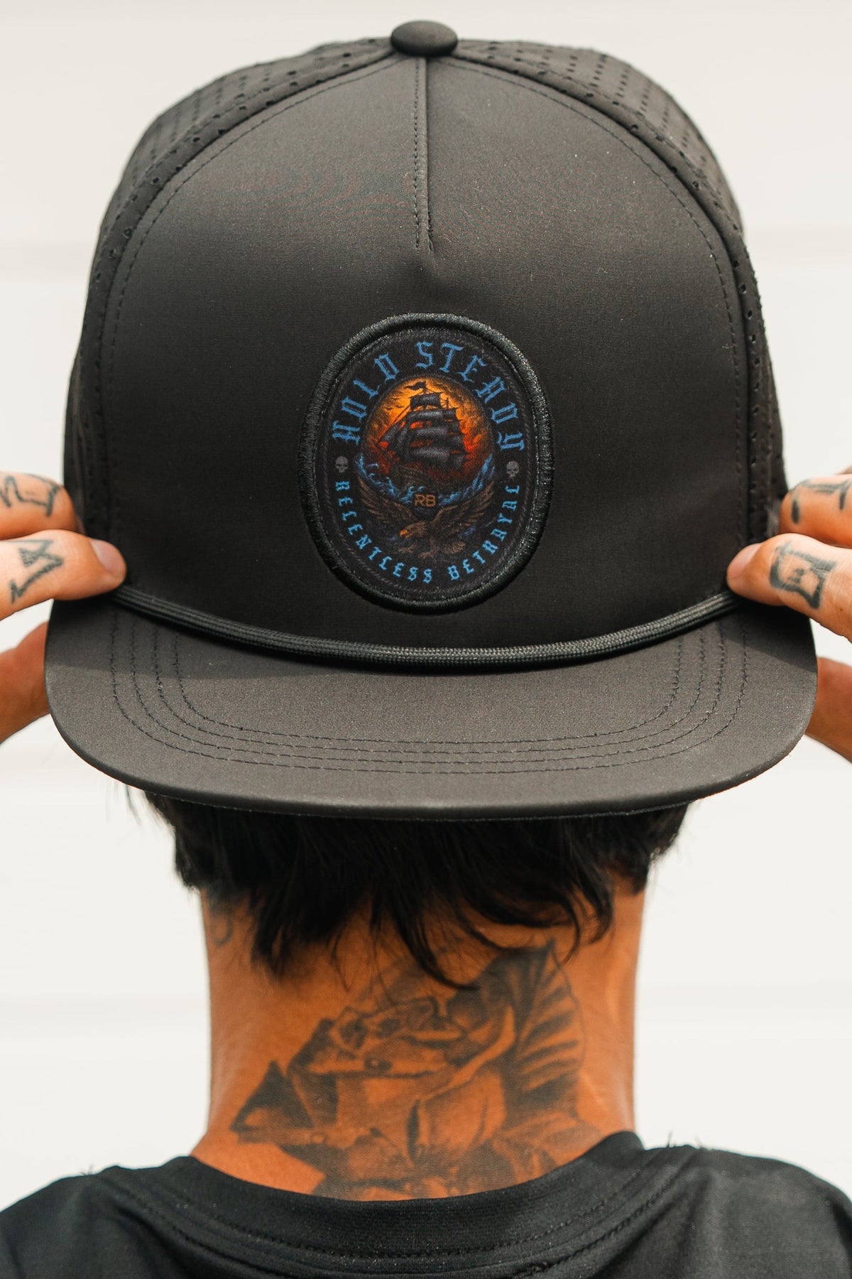 A person adjusts the brim of the Relentless Betrayal Hold Steady Snapback, featuring a circular patch and water-resistant design. Visible finger tattoos and a large floral neck tattoo stand out against a plain, light-colored background.