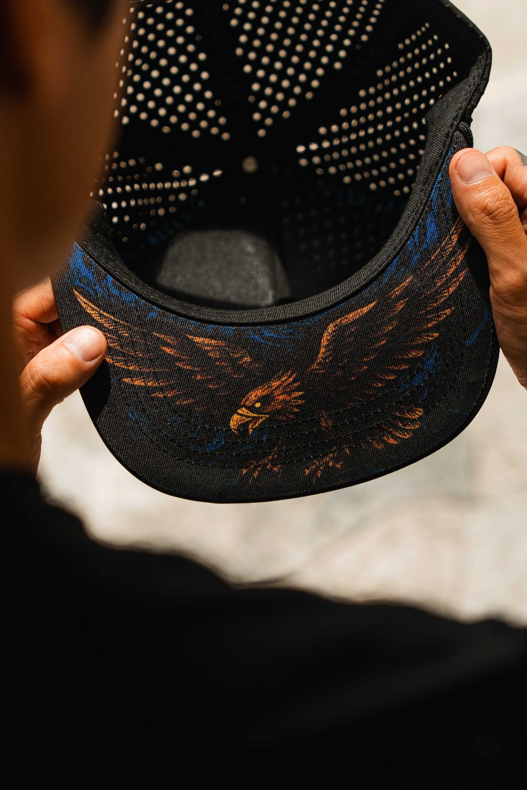 A person holds a black Relentless Betrayal Hold Steady Snapback, viewed from behind to reveal the colorful eagle illustration with outstretched wings on the underside of the premium hat’s brim.
