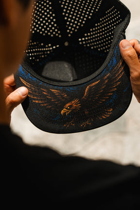 A person holds a black Relentless Betrayal Hold Steady Snapback, viewed from behind to reveal the colorful eagle illustration with outstretched wings on the underside of the premium hat’s brim.