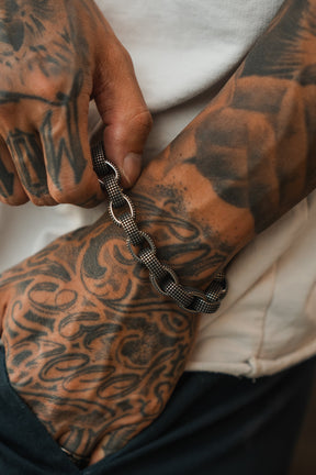 A tattooed hand wears the Relentless Betrayal Seeker Bracelet—a chunky, hand-forged stainless steel chain made in the USA—resting casually in a dark pant pocket and paired with a crisp white shirt.