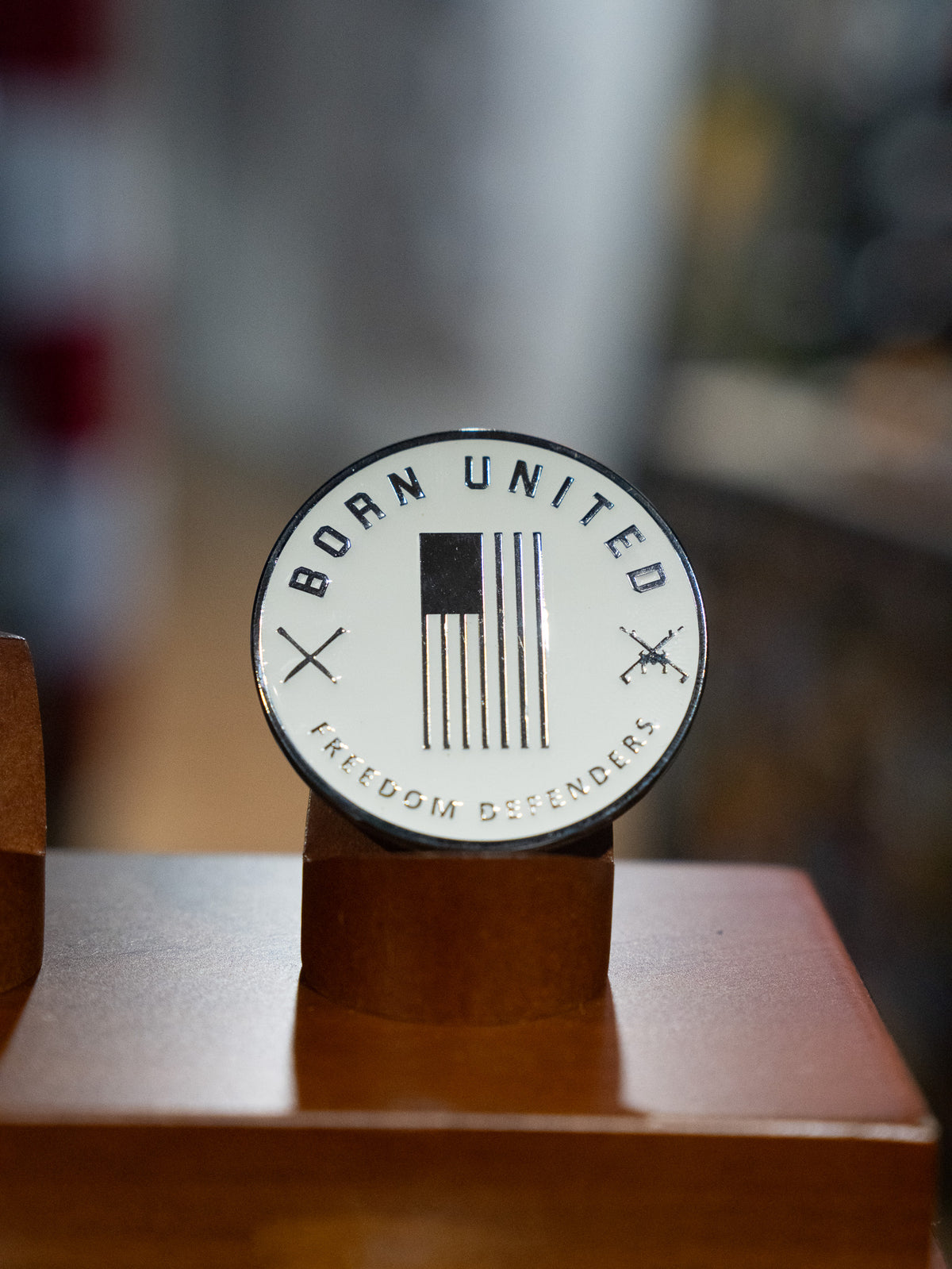 The Last Laughs Club Challenge Coin by Born United Private Label features a black and white US flag, crossed rifles, and the words “BORN UNITED” and “FREEDOM DEFENDERS”—a bold display piece on its wooden stand for any member.