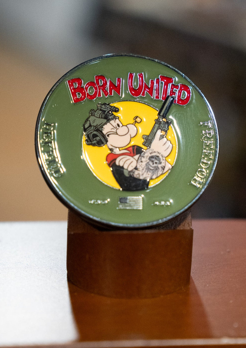 The Sailor Man Challenge Coin by Born United Private Label is a round green coin with Popeye holding a rifle, wearing a military helmet and tattoos, plus "Born United" in red at the top and "Freedom" and "Justice" along the sides.