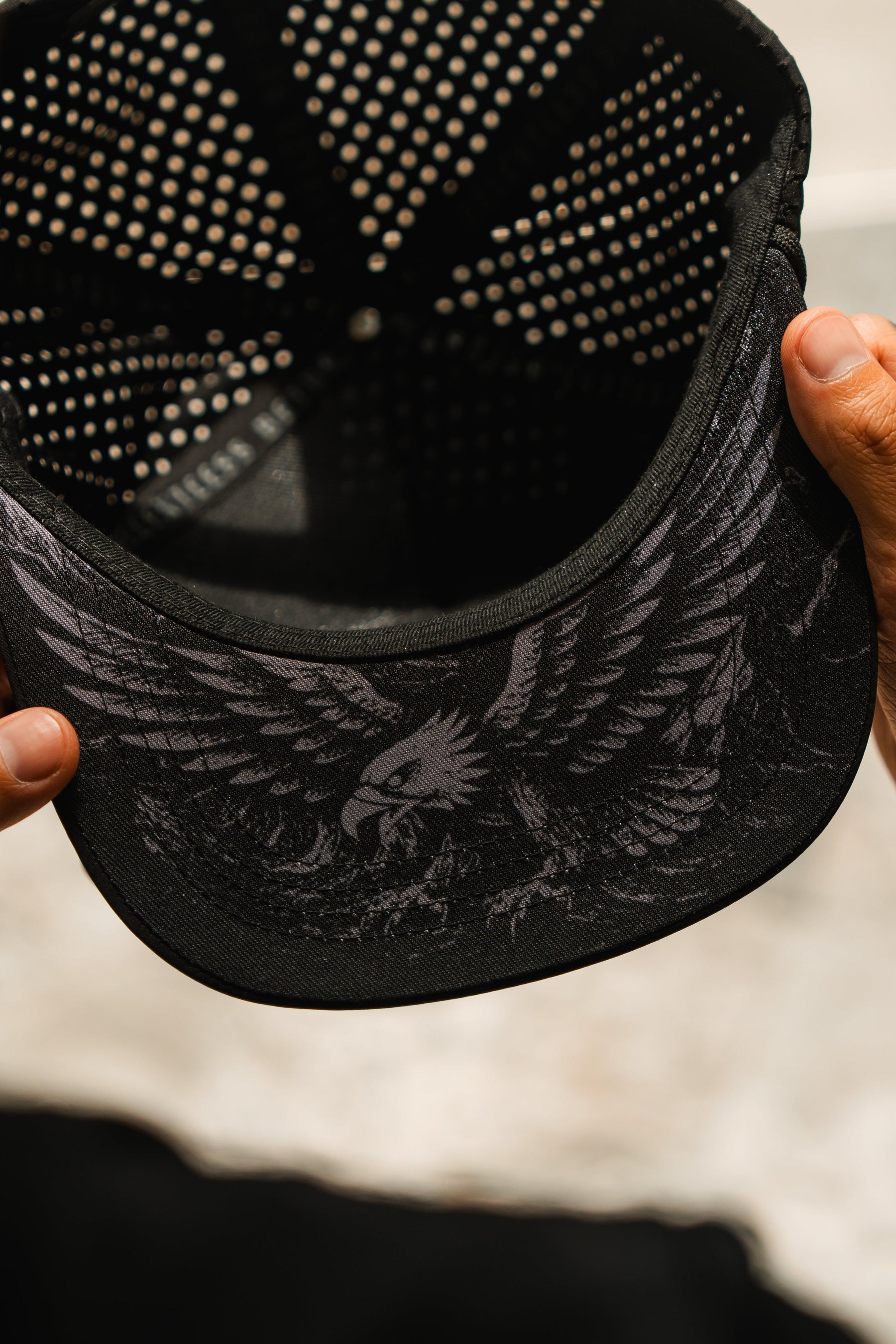 A close-up of hands holding the Relentless Betrayal Hold Steady Ghost Snapback, revealing the eagle illustration under the brim. This water-resistant premium hat features a perforated inner lining for comfort.