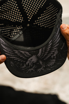 A close-up of hands holding the Relentless Betrayal Hold Steady Ghost Snapback, revealing the eagle illustration under the brim. This water-resistant premium hat features a perforated inner lining for comfort.