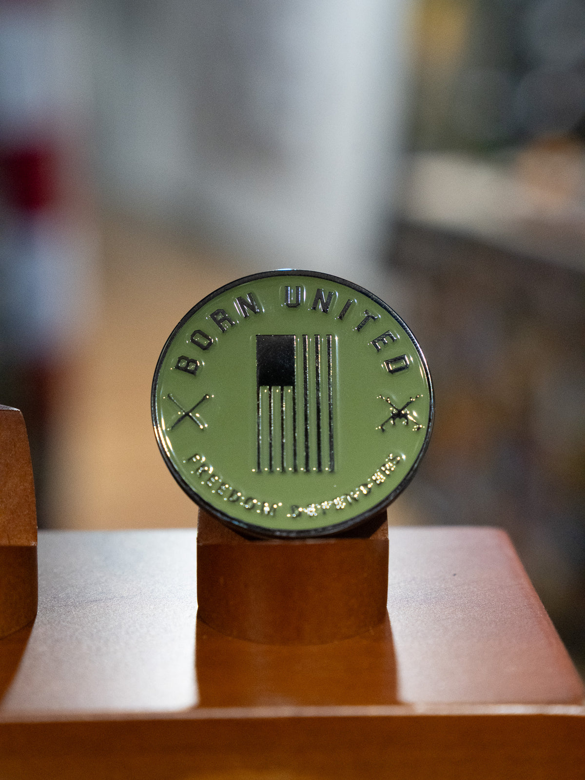 The Sailor Man Challenge Coin by Born United Private Label features a round, olive-green design with a U.S. flag, crossed rifles, and “BORN UNITED” inscribed on top. The coin is displayed upright on a brown stand against a blurred background.