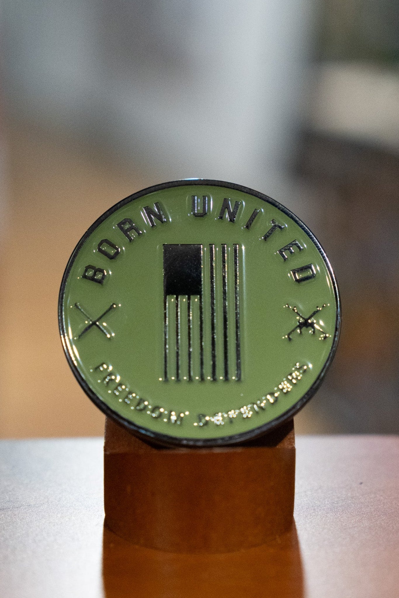 The Sailor Man Challenge Coin by Born United Private Label features a round, olive-green design with a U.S. flag, crossed rifles, and “BORN UNITED” inscribed on top. The coin is displayed upright on a brown stand against a blurred background.
