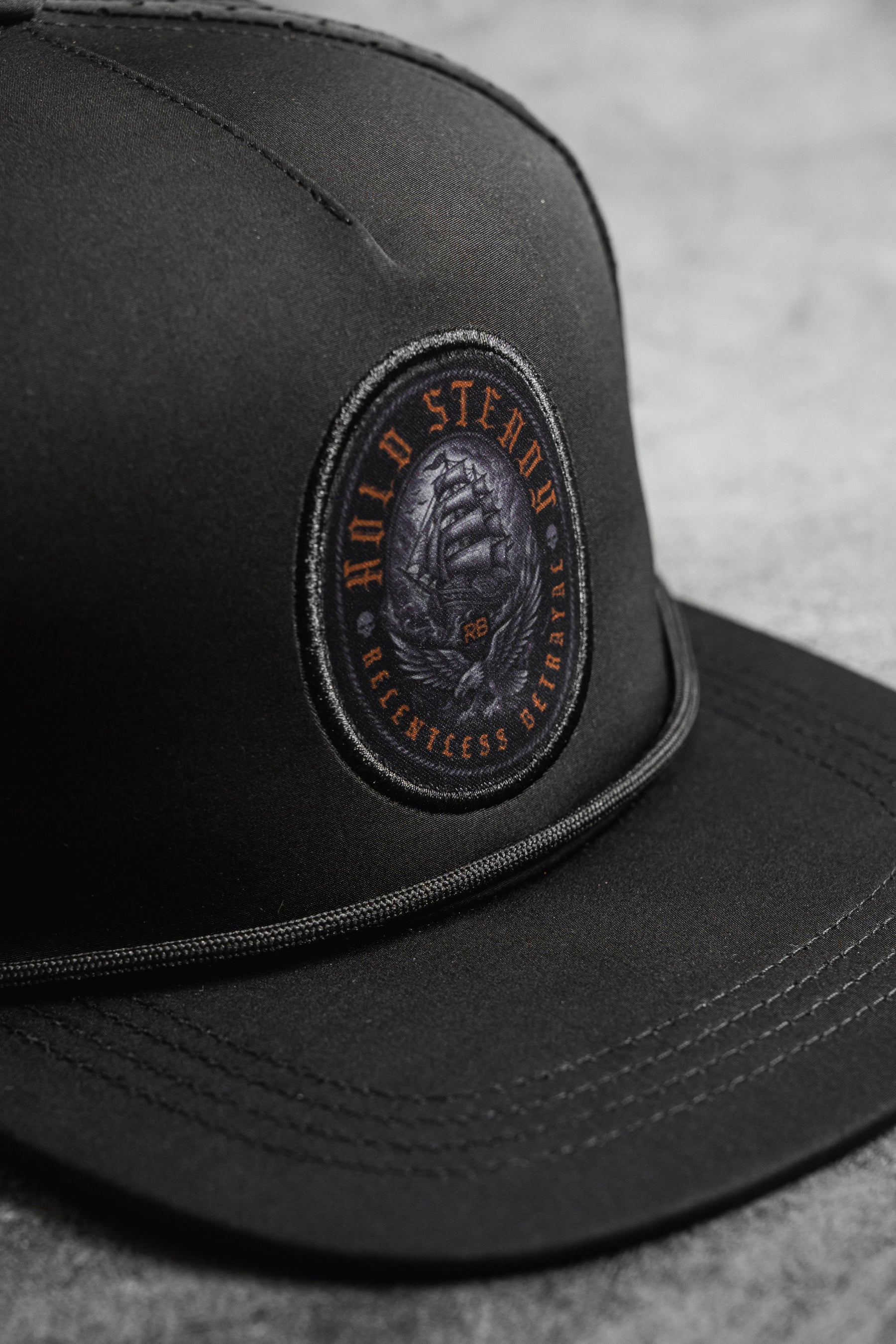 A close-up of the Relentless Betrayal Hold Steady Ghost Snapback, a black cap with a circular patch reading "WILD STEADS RELENTLESS ETERNAL" and a hand gripping stone, displayed on a gray surface.