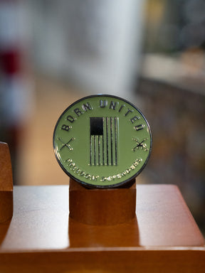 The Sailor Man Challenge Coin by Born United Private Label features a round, olive-green design with a U.S. flag, crossed rifles, and “BORN UNITED” inscribed on top. The coin is displayed upright on a brown stand against a blurred background.