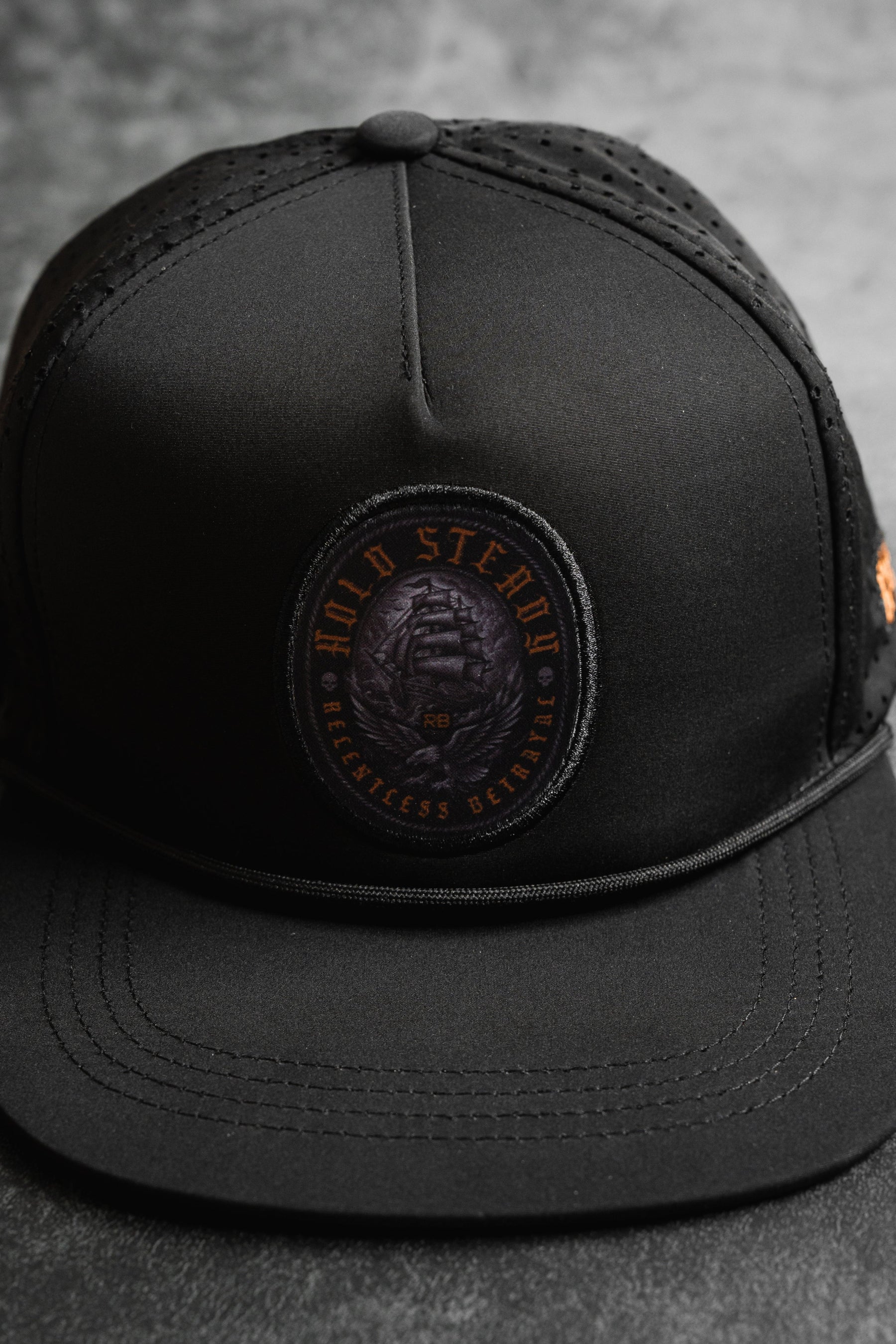 A close-up of the Hold Steady Ghost Snapback by Relentless Betrayal—a premium snapback baseball cap with a round patch reading HOLD STEADY BREATHLESS BETRAYED in orange and showcasing a coiled snake.