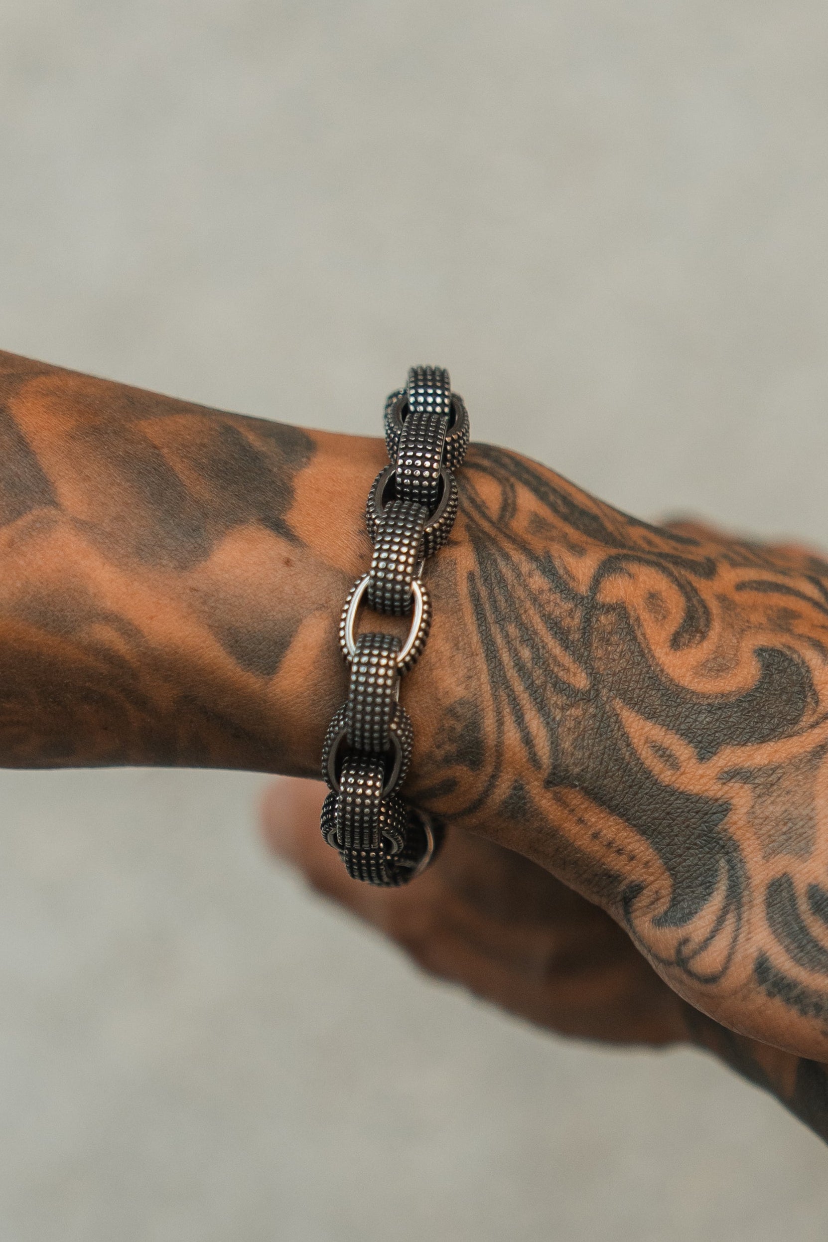 A tattooed arm wears the Relentless Betrayal Seeker Bracelet, a hand-forged stainless steel black and silver chain-link design, shown close-up against a neutral background.