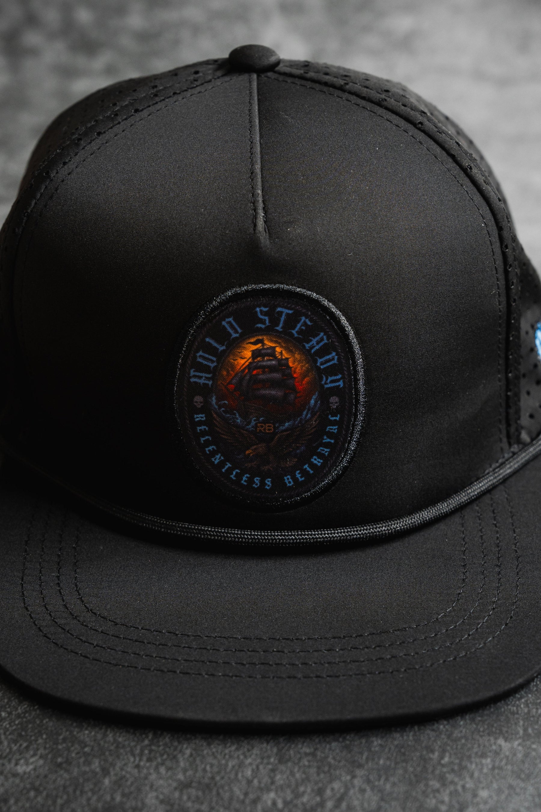 The Relentless Betrayal Hold Steady Snapback is a black baseball cap with a circular ship patch and "Hold Steady, Relentless, and Reliable" embroidered around the edge.