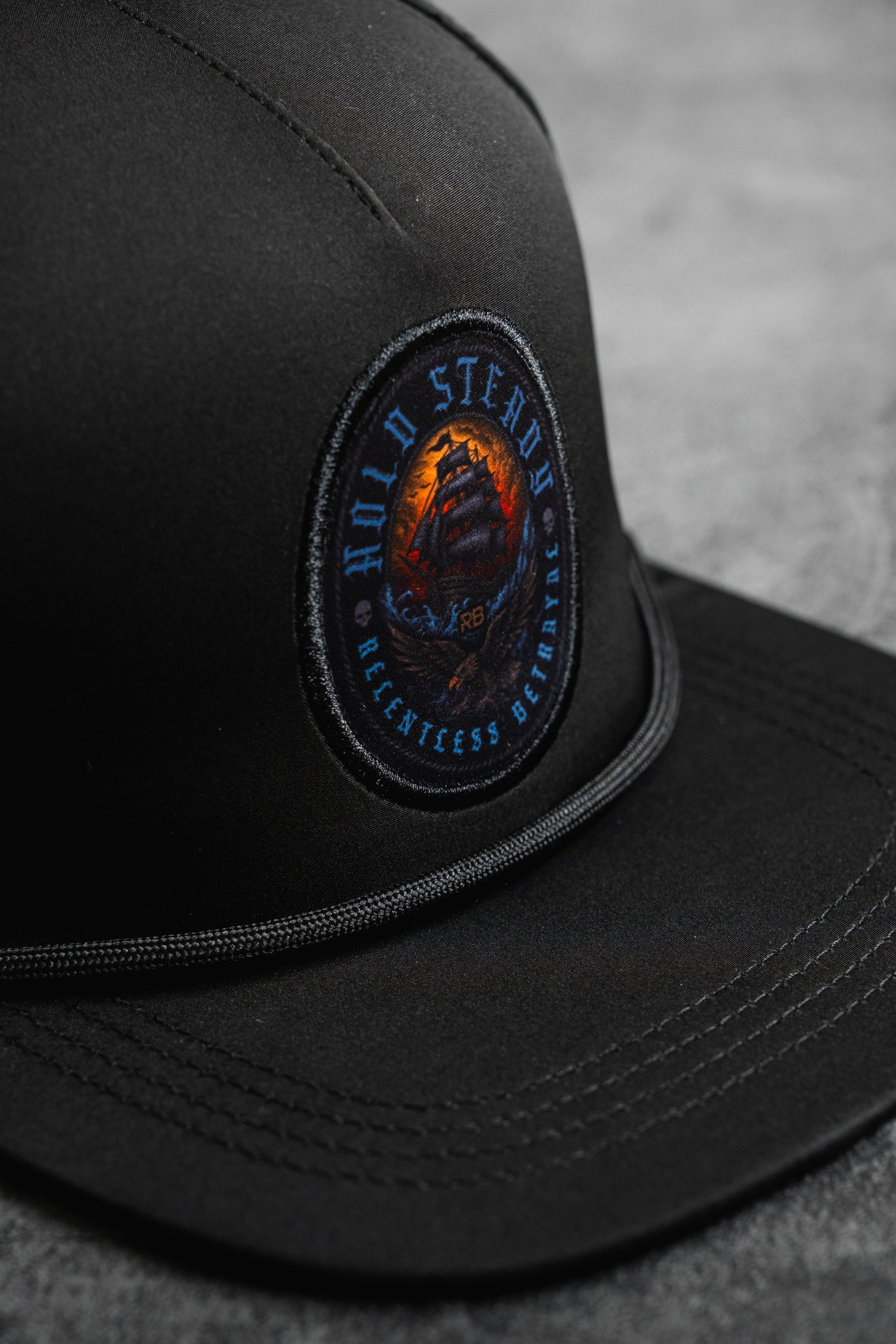 A close-up of the Hold Steady Snapback by Relentless Betrayal, featuring a round embroidered ship patch with "HOLD STEADY" and "RELENTLESS BELIEFING." This water-resistant hat rests on a gray surface.