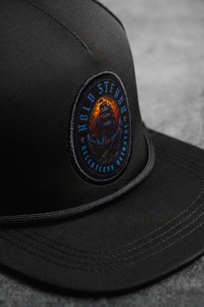 A close-up of the Hold Steady Snapback by Relentless Betrayal, featuring a round embroidered ship patch with "HOLD STEADY" and "RELENTLESS BELIEFING." This water-resistant hat rests on a gray surface.